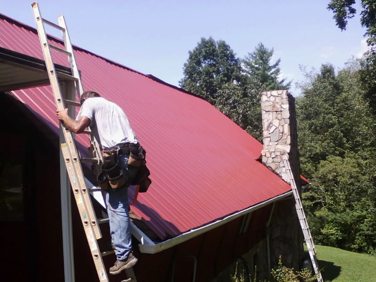 Roofer on a ladder beside a red metal roof and stone chimney — Soffit & Fascia Repair by Roofing Brothers in Mossyrock